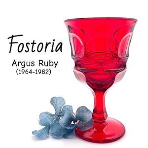 Puede incluir: Una copa de cristal Fostoria Argus de color rojo rubí. El cristal tiene un diseño texturizado y una base redonda. El texto "Fostoria Argus Ruby (1964-1982)" se muestra en negro. Un racimo de flores azules está en la base del cristal.