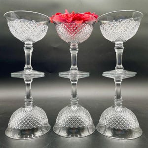 Könnte beinhalten: Drei klare Kristall-Champagner-Coupe-Gläser mit Diamantschliffmustern. Ein Glas enthält eine rote Rose. Die Gläser sind in einer Reihe angeordnet, wobei die Basen nach oben zeigen.