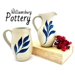 May include: Two off-white ceramic pitchers with blue leaf patterns and handles. The text Williamsburg Pottery is printed in black above the pitchers. A cluster of red flowers sits next to the taller pitcher on a thick, light-colored book.