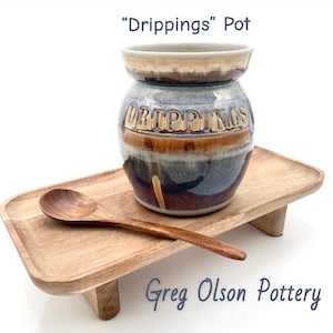 May include: A handcrafted ceramic "Drippings" pot with a wooden spoon on a wooden tray. The pot is glazed in shades of blue, brown, and beige, with the word "DRIPPINGS" embossed on the front. The tray and spoon are made of light-coloured wood.