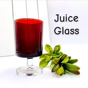 Puede incluir: Un vaso de zumo rojo con tallo y base transparentes, con las palabras "Juice Glass" en negro. El vaso está junto a una ramita de plantas suculentas verdes. El vaso está sobre una superficie blanca.