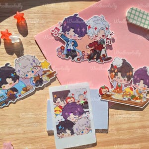 May include: A collection of colorful anime-style stickers featuring various characters in different poses. The stickers depict characters with purple, white, and black hair, some holding microphones or food. The stickers are on a wooden surface and pink paper.
