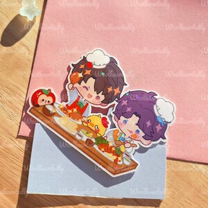 May include: A colorful sticker featuring two cartoon chefs with food on a wooden serving board. The sticker has a light blue background and is placed on a pink envelope. The text "Woolheartedly" is visible.