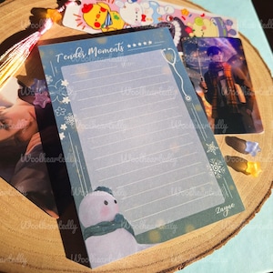 以下が含まれることがあります： 雪だるまの絵と「Tender Moments」という文字が書かれた、青と緑の罫線入りのメモ帳。メモ帳には、書き込み用の白い紙が挟まれています。