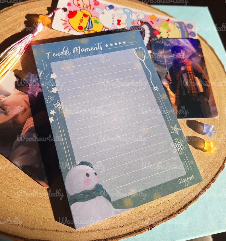 以下が含まれることがあります： 雪だるまの絵と「Tender Moments」という文字が書かれた、青と緑の罫線入りのメモ帳。メモ帳には、書き込み用の白い紙が挟まれています。