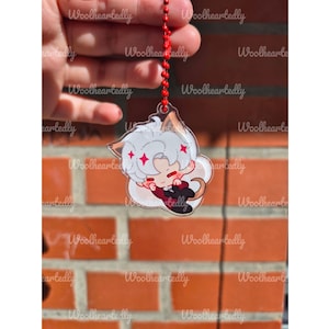 Pode incluir: Um pingente de acr&iacute;lico transparente com um personagem estilo chibi de cabelo branco, orelhas pontudas e cauda, vestindo um terno escuro com gravata vermelha. O pingente est&aacute; preso a uma corrente de bolinhas vermelha com detalhes de estrelas.