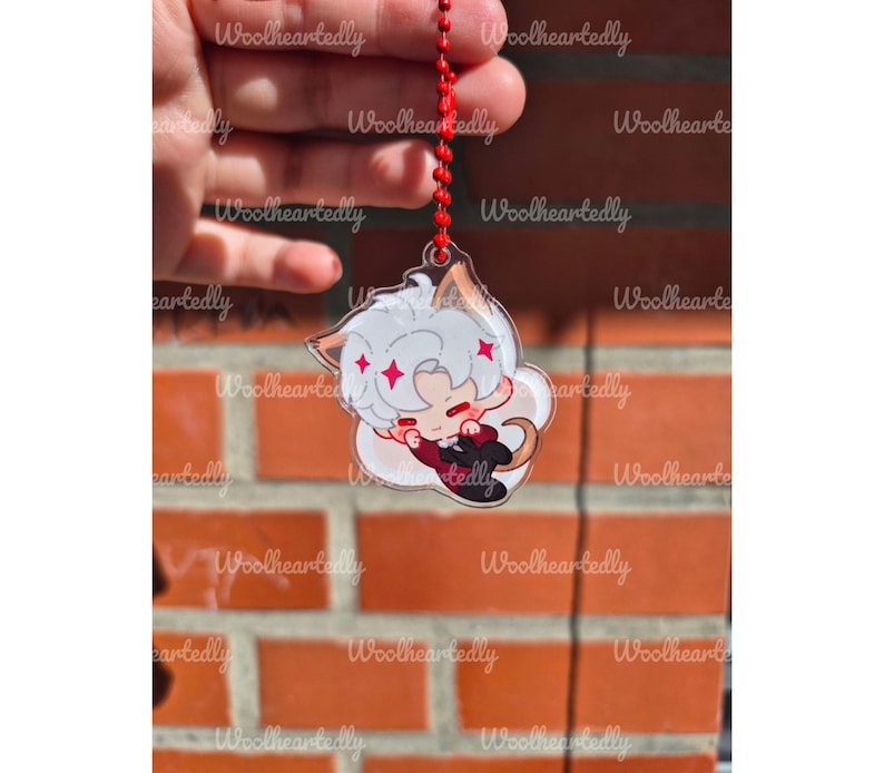 Pode incluir: Um pingente de acr&iacute;lico transparente com um personagem estilo chibi de cabelo branco, orelhas pontudas e cauda, vestindo um terno escuro com gravata vermelha. O pingente est&aacute; preso a uma corrente de bolinhas vermelha com detalhes de estrelas.