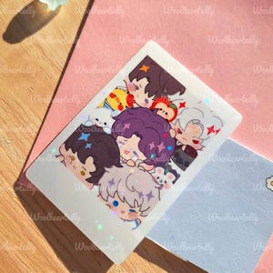 May include: A holographic sticker featuring a group of cartoon characters with various hair colors and expressions. The sticker is rectangular with rounded corners and has a shiny, iridescent finish. The background includes a pink card, a light blue card, and a wooden surface.
