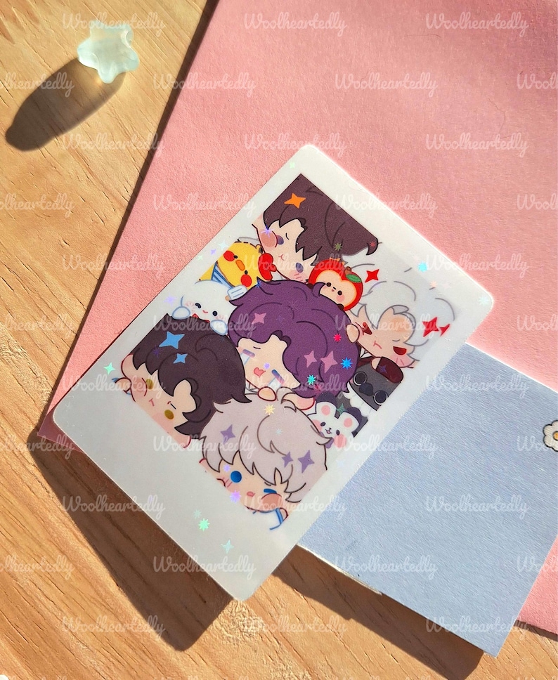 May include: A holographic sticker featuring a group of cartoon characters with various hair colors and expressions. The sticker is rectangular with rounded corners and has a shiny, iridescent finish. The background includes a pink card, a light blue card, and a wooden surface.