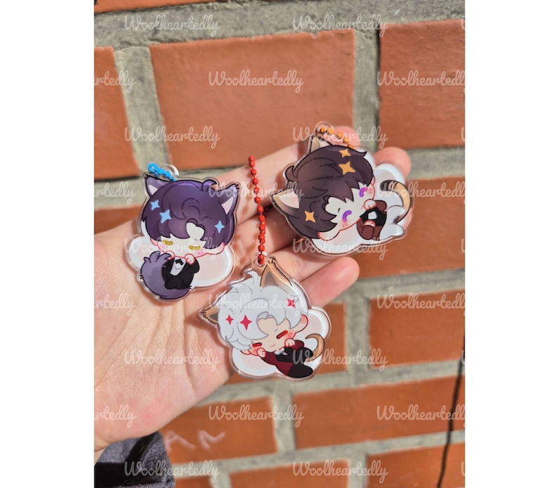 Pode incluir: Tr&ecirc;s pingentes de acr&iacute;lico transparente com personagens estilizados de anime com orelhas de gato. Um personagem tem cabelo escuro, um tem cabelo claro e um tem cabelo escuro com um la&ccedil;o azul. Cada pingente est&aacute; preso a uma corrente de bolinhas vermelha.
