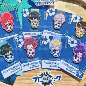 Peut inclure: Ensemble de marque-pages magnétiques avec des personnages de style anime tenant des ballons de football. Chaque marque-page présente un personnage différent avec des couleurs de cheveux et des motifs uniques, sur fond bleu. Le texte "Reading Companion Bookmark" est visible.