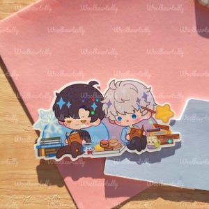 May include: A sticker featuring two anime-style characters, one with dark hair and the other with light hair, both reading books. The sticker includes stacks of books, pastries, and a star-shaped wand. The background is a pink and blue surface.