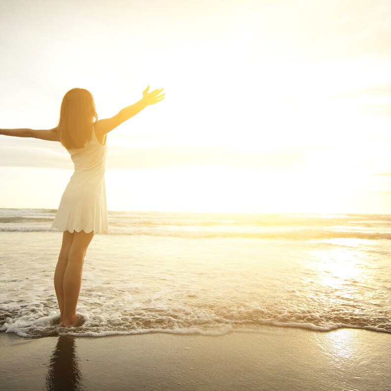 May include: A woman in a white dress stands on a sandy beach with her arms outstretched, facing the setting sun. The ocean waves lap at her feet.