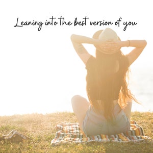 May include: A woman wearing a straw hat sits on a blanket with her arms raised above her head. The text "Leaning into the best version of you" is written above her.