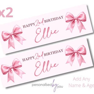 May include: Two light pink birthday banners with the text "Happy 2nd Birthday Ellie" and pink bow illustrations. The text is in a darker pink. The banners also include the text "Add Any Name & Age" and the words "personalised by you".