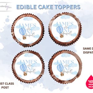 May include: Four edible cake toppers with a blue hot air balloon design, clouds, and the text "JAMES is ONEderful!". The toppers have a chocolate-coloured border. The image also includes text that says "EDIBLE CAKE TOPPERS".