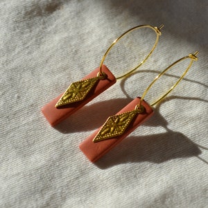 May include: A pair of gold hoop earrings with a coral-coloured rectangular pendant featuring a gold diamond-shaped charm.
