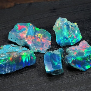 Puede incluir: Cinco piezas de ópalo en bruto, con una base azul verdosa y destellos iridiscentes de color, están dispuestas sobre una superficie negra.