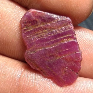 Könnte beinhalten: Ein tiefroter Rubin-Edelstein mit einer rauen, facettierten Oberfläche. Der Rubin hat ein gestreiftes Muster und ist ein natürlicher, unpolierter Stein.