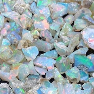 Może przedstawiać: Widok z bliska na stos małych, opalizujących kawałków opalu. Kawałki są w różnych odcieniach niebieskiego, zielonego i białego, z mieniącym się, tęczowym efektem. Tekst "opal factory" jest widoczny w lewym dolnym rogu obrazu.