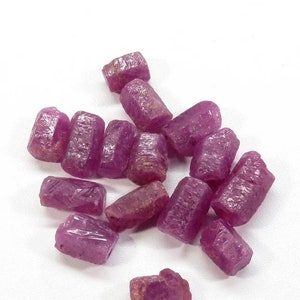 Puede incluir: Una colección de gemas de rubí en bruto sin cortar. Las gemas son de un color rojo rosado intenso, con superficies rugosas y texturizadas. Las piedras tienen formas irregulares y un aspecto natural sin pulir. Los rubíes están dispersos sobre una superficie blanca.