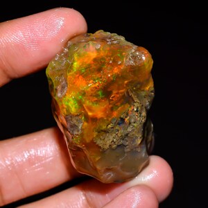 Peut inclure: Une pierre d'opale brute et non taillée aux couleurs vives. L'opale présente un mélange de teintes orange, jaune et verte, avec une surface rugueuse et texturée. La pierre est tenue dans une main sur fond sombre.
