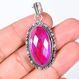 May include: A silver pendant with an oval, faceted, magenta gemstone. The gemstone is set in a decorative silver frame with a textured border. The pendant has a silver bail for attaching to a chain.