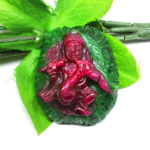 May include: A carved gemstone pendant featuring a deep red figure playing a flute, set against a textured dark green oval background. The central figure is depicted in a seated position with detailed facial features and traditional headwear.