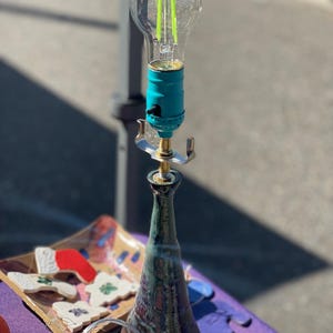 May include: A ceramic table lamp with a teal blue base and a clear glass shade with a green LED light bulb. The lamp is sitting on a purple table with a ceramic tray of decorative ornaments.