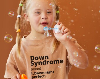 Pode incluir: Uma menina jovem com tranças loiras sopra bolhas com uma varinha de bolhas azul e branca. Ela está vestindo uma camiseta marrom com o texto "Down Syndrome (noun) 1. Down right perfect."