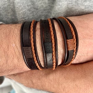 May include: A multi-layered bracelet set featuring dark brown leather bands with braided tan accents. One band is embossed with the words "Forever Always". The bracelets are stacked on top of each other.