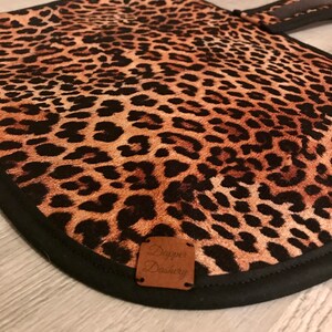 Puede incluir: Un guante de cocina con estampado de leopardo, borde negro y asa a juego. El guante presenta una etiqueta de cuero marr&oacute;n con el texto "Dapper Dashery".