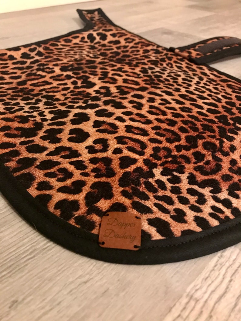 Puede incluir: Un guante de cocina con estampado de leopardo, borde negro y asa a juego. El guante presenta una etiqueta de cuero marr&oacute;n con el texto "Dapper Dashery".