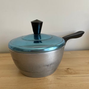 May include: A small, vintage saucepan with a light blue lid and a black handle. The pot is silver-coloured with a black knob on the lid. The handle is curved and attached to the side. The saucepan is likely made of metal.