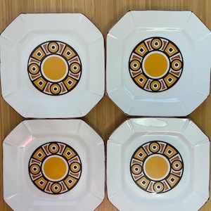 May include: Four white octagonal plates with a retro design. Each plate features a central yellow circle surrounded by concentric rings of brown, orange, and black geometric patterns. The plates have a brown trim.