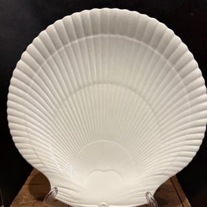 May include: A white ceramic dish shaped like a large scallop shell. The surface features raised, radiating ridges that extend from the base to the scalloped edge, creating a textured, fan-like appearance with a glossy finish.