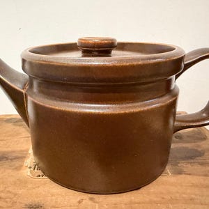 May include: A solid brown ceramic teapot featuring a cylindrical body, a wide flat lid with a central knob, a curved handle on the right side, and a short, angled spout on the left. The teapot has a smooth, uniform matte finish throughout.