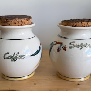 May include: A white ceramic mug with a black handle and the text "But First, Coffee" in a bold, black font. The mug is set against a blurred background, suggesting a cosy setting.