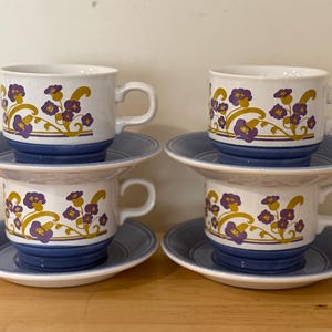 May include: Four matching teacups and saucers. The cups are white with a blue base and handle, and feature a floral design in purple and gold. The saucers are light blue. The set is arranged on a wooden surface.