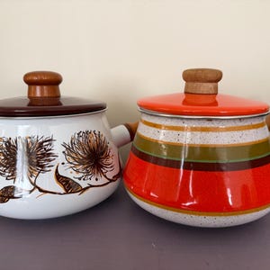 May include: Two vintage enamel pots with wooden handles and lids. One pot is white with brown floral designs, the other is orange with green and brown stripes. Both have wooden knobs.