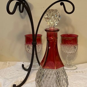 May include: A clear glass decanter with a red base and a diamond pattern. The decanter is topped with a clear glass stopper with a diamond pattern. The decanter is sitting on a white tablecloth with a black metal candelabra behind it. Two clear glass goblets with red bases and diamond patterns are on either side of the decanter.