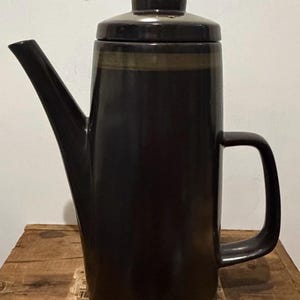 May include: A tall, cylindrical ceramic coffee pot in a deep, dark brown glaze. It features a long, angled spout, a matching lid with a small knob handle, and a sturdy, rectangular side handle. A subtle, lighter brown band encircles the upper rim.