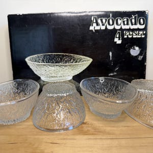 May include: A set of four clear glass bowls with a textured surface, arranged in front of a black box with the text "Avocado 4 PC SET". The bowls have an oval and round shape.