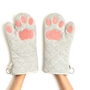 May include: A pair of gray oven mitts with pink paw print designs. The mitts are quilted and have a hanging loop.