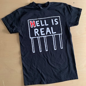 May include: Black t-shirt with a white graphic of a sign that reads "HELL IS REAL" in white text with the word "HELL" in red. The sign has white vertical lines below it.