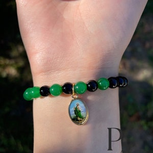 May include: A green and black beaded bracelet with a gold charm featuring a religious image. The charm is oval shaped and has a picture of a man in a green robe. The bracelet is on a person's wrist.
