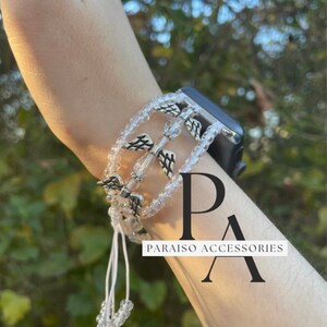 May include: A white beaded bracelet with silver angel wing charms. The bracelet is worn on a wrist and has a white string attached to it. The text "Paraiso Accessories" is visible in the image.