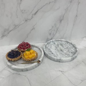 May include: Two round white marble serving trays with a light gray veining pattern. One tray has three mini tarts with fruit toppings. The tarts are filled with blueberries, raspberries, and diced mango.