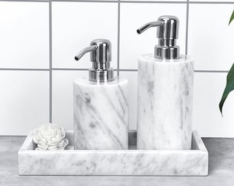 May include: A marble bathroom set with two soap dispensers and a rectangular tray. The dispensers have silver pumps and a white and grey veined pattern. A decorative white flower sits on the tray. The set is on a grey surface.