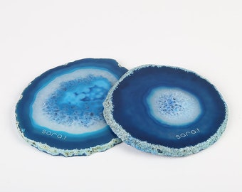 Op de afbeelding: Twee blauwe agaat onderzetters met witte en lichtblauwe centra. De onderzetters hebben een natuurlijke, organische vorm en een gepolijst oppervlak. De tekst "sara.l" is op het oppervlak van elke onderzetter gedrukt.
